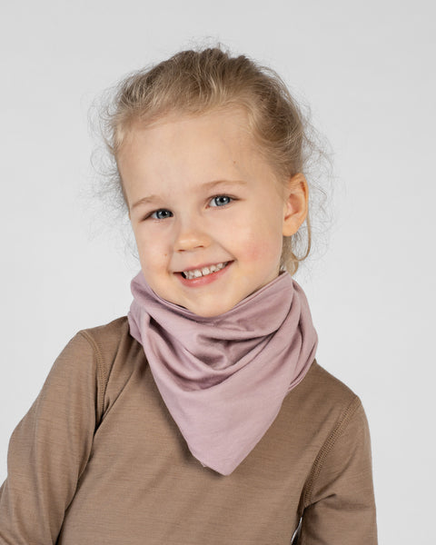 A studio portrait of a young blonde girl looking directly at the camera with a big smile. She is wearing a dusty pink bandana tied around her neck with the point facing down, over a light brown long-sleeved shirt. The background is a plain light grey.