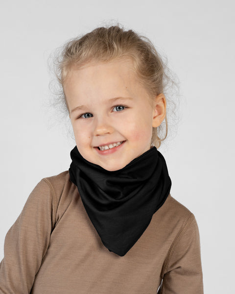 A studio portrait of a young blonde girl looking directly at the camera with a big smile. She is wearing a solid black bandana tied around her neck with the point facing down, over a light brown long-sleeved shirt. The background is a plain light grey.