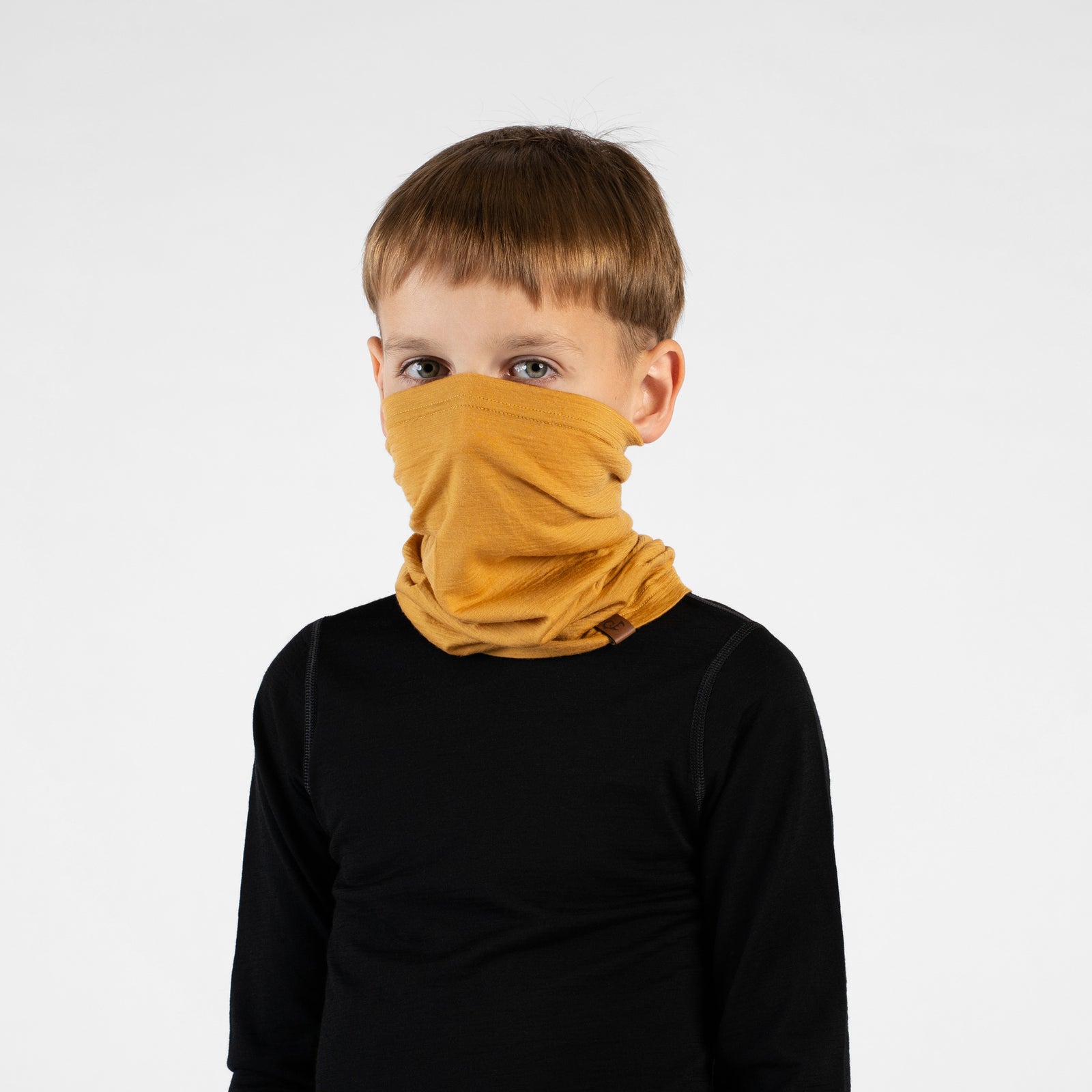 A close-up portrait of a young boy looking off to his left in a three-quarter profile against a white background. He is wearing a black long-sleeved top and a mustard yellow neck gaiter pulled up over his nose and mouth, showing how it bunches comfortably around his neck.