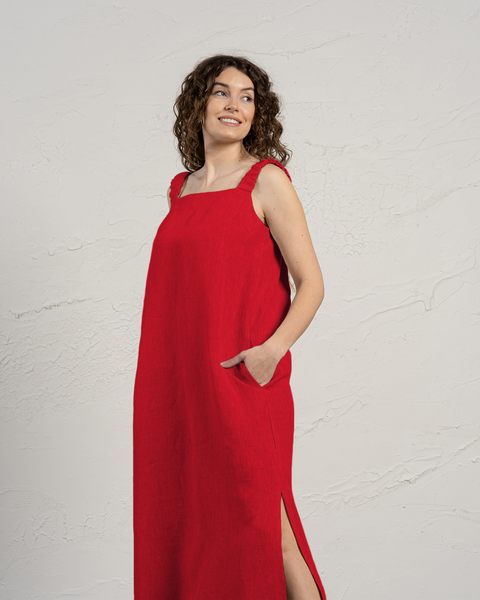 A woman with curly brown hair is standing in front of a textured off-white wall. She is wearing a sleeveless, linen floor-length pure red dress with wide straps and a square neckline. One of her hands is tucked into the side pocket of the dress, and there is a slit visible at the bottom of the dress on one side. She is looking towards the upper left corner of the frame with a smile. The lighting is even, casting soft shadows.