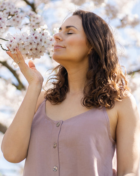 A close-up shot shows a woman with long, dark, wavy hair, wearing a faded rose sleeveless dress, with her eyes closed and a serene expression. She holds a sprig of white blossoms up to her nose, appearing to be smelling them. The background is blurred, showing more white blossoms and a bright sky, suggesting an outdoor setting on a sunny day.