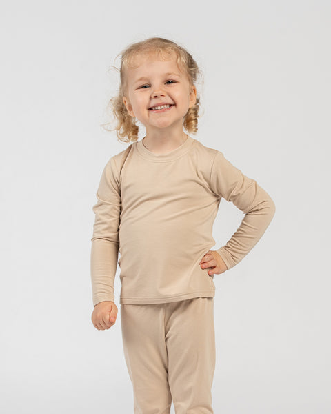 Happy child standing in beige bamboo long-sleeve top and bottoms with one hand on hip.