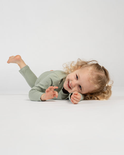 Child in green long-sleeve outfit lying playfully on the floor, smiling at the camera with one leg raised and curly hair spread out on the ground against a plain background.