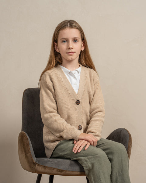 A girl is sitting on a gray chair, with her hands put neatly on her knees. She is wearing the menique kids' merino wool cardigan in beige color. The picture emphasizes the v-neck shape of the cardigan.
