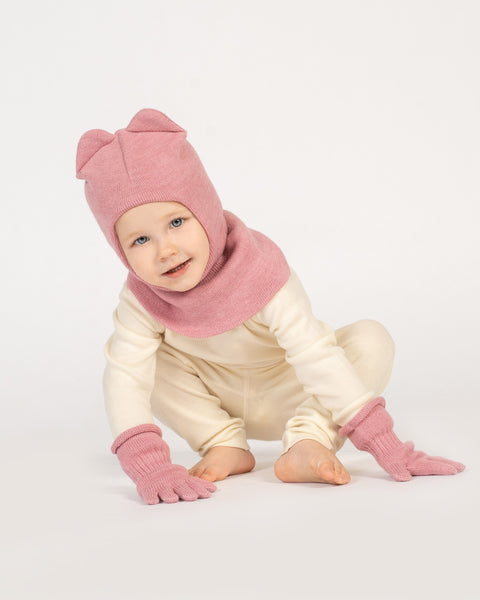 A young child with blue eyes and a wide, happy smile. They are wearing a pink balaclava with ears, matching pink mittens, and a light-colored long-sleeved shirt and pants.
