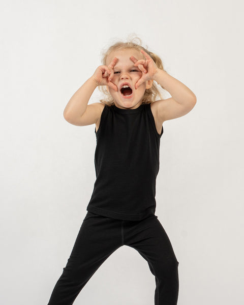 A child with curly blonde hair stands against a white background, wearing a black sleeveless top and black pants. The child is looking at the camera with their mouth wide open and both hands raised to their eyes, forming a shape like glasses or binoculars with their fingers.