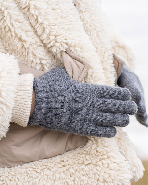 A person wearing a cream-colored sherpa coat with beige pockets and a light-colored ribbed cuff is putting on or adjusting a dark grey knitted glove. The background is a blurred, light outdoor scene.