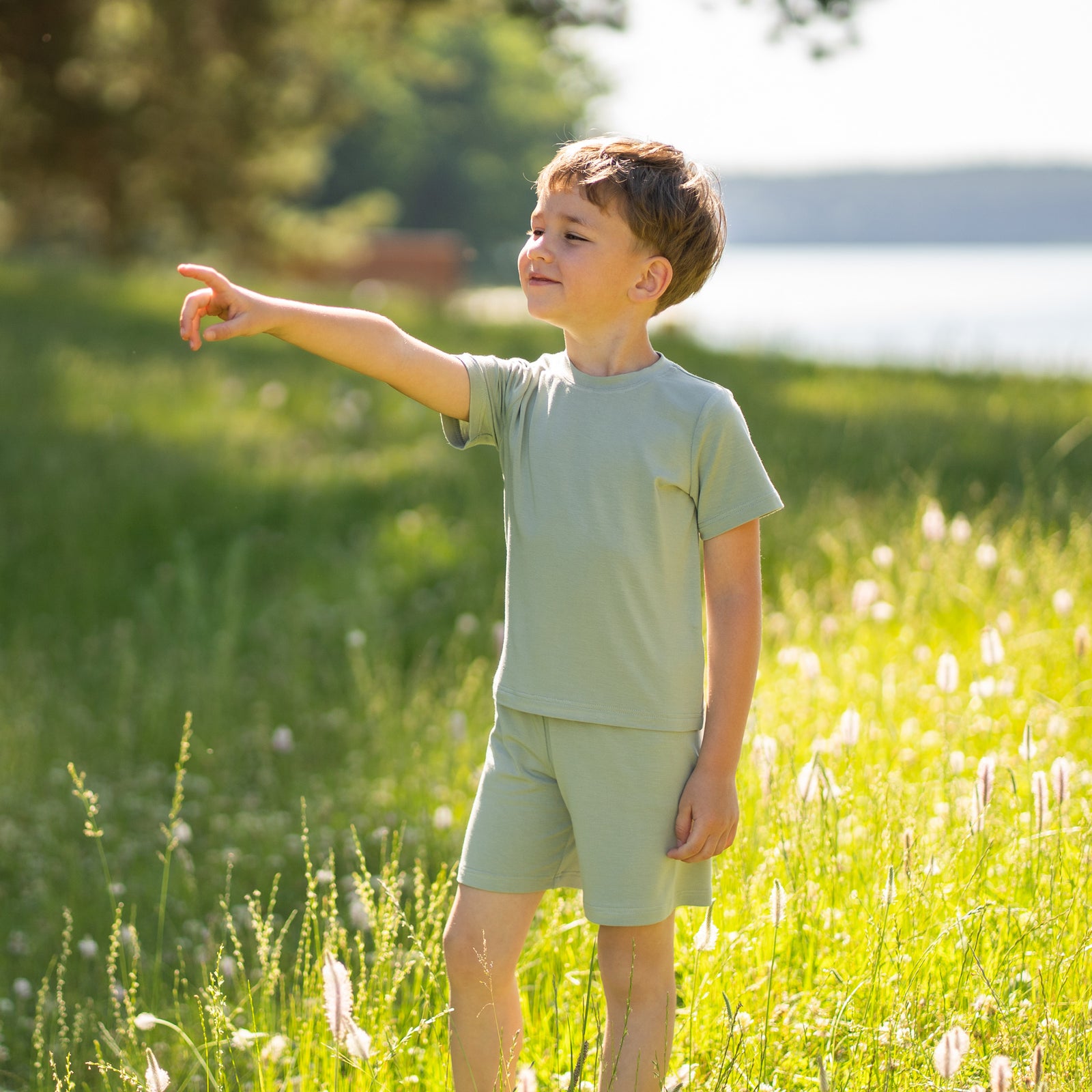 A studio shot, cropped from the chest down, of a young child standing barefoot. The child has their hands resting on their hips. They are wearing a matching set of a light sage green short-sleeved t-shirt and shorts. The background is a plain light grey.