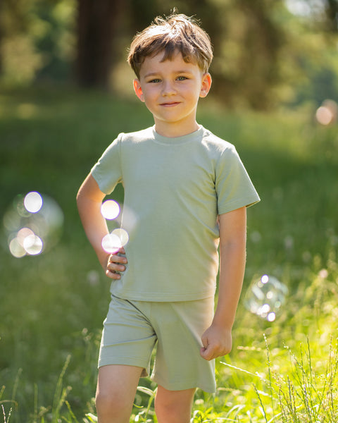 Young boy in a sunny meadow wearing a light green bamboo T-shirt and matching shorts. The lightweight kids' outfit is showcased in a natural outdoor setting, highlighting its soft, breathable comfort for everyday adventures.