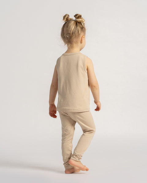 Little girl in a beige sleeveless top and leggings set seen from the back, walking barefoot on a light background.