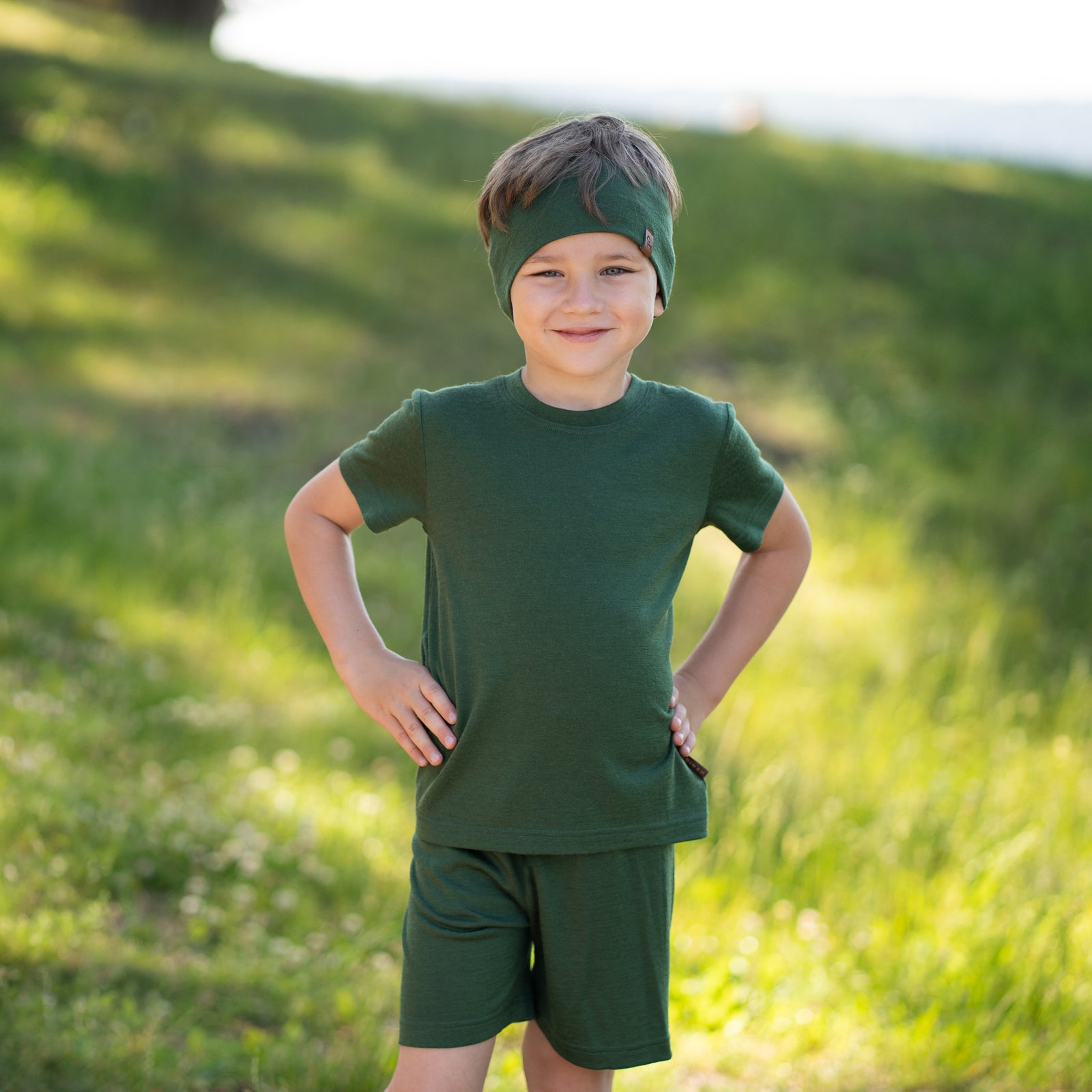 A close-up studio portrait of a smiling young child looking up and to the left against a plain light grey background. She is wearing a black short-sleeved t-shirt and a wide dark green headband with a small brown logo tag on the side, her hair pulled back in a ponytail.