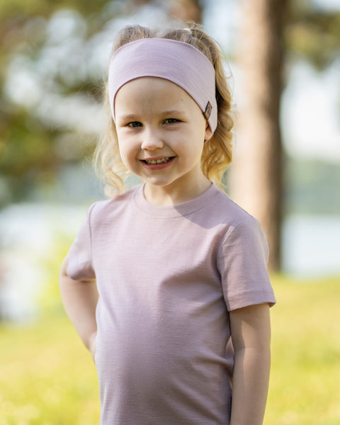 Young girl smiling outdoors wearing a dusty pink Merino wool T-shirt and a matching Merino wool headband. The soft, breathable accessories and clothing are showcased in a sunny natural setting, offering comfortable everyday wear for active ki