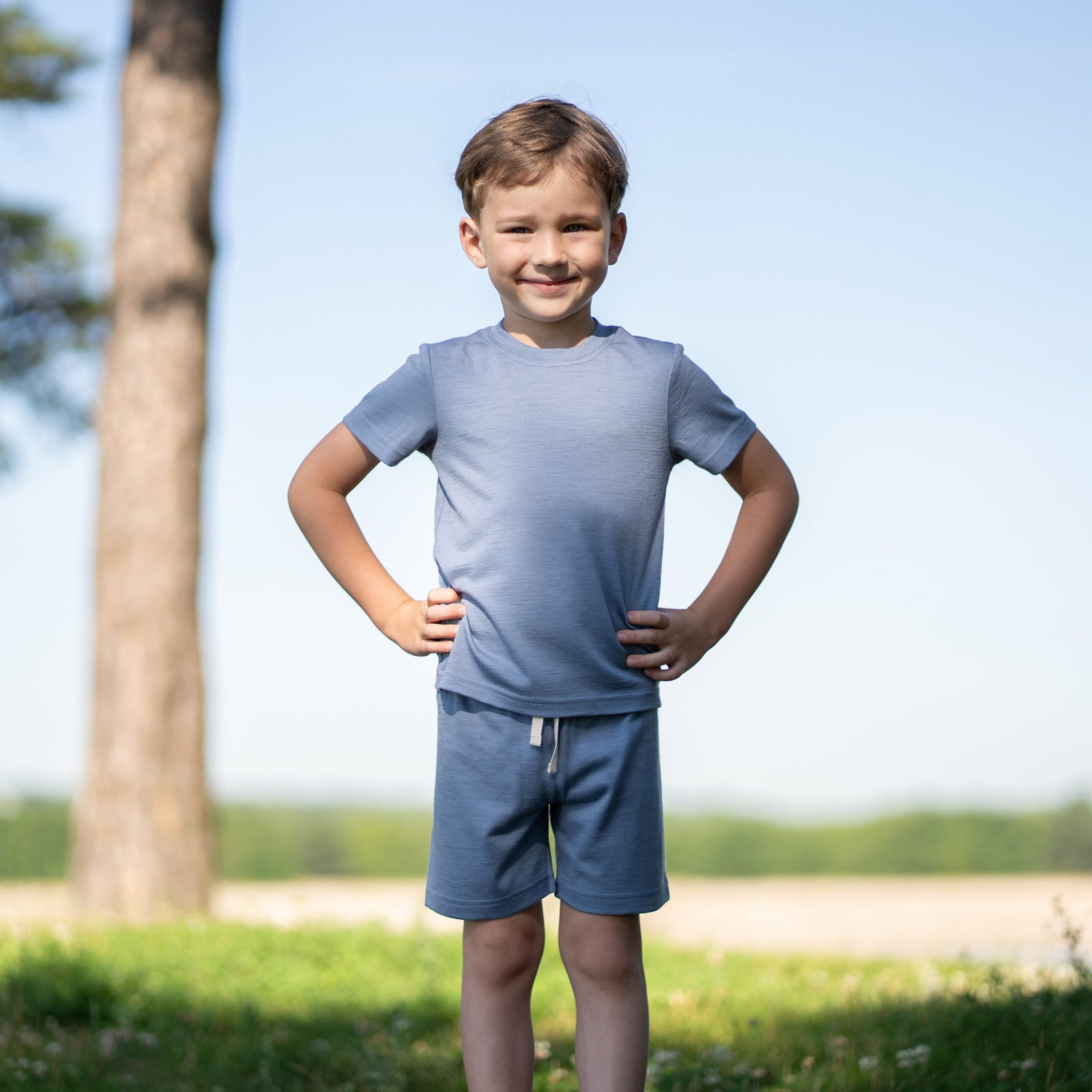 A young girl with blonde hair styled in two pigtails smiles at the camera. She is wearing a dusty blue merino short-sleeve t-shirt, matching dusty blue merino shorts, and a matching dusty blue merino ribbon headband. She is standing against a plain light gray background.