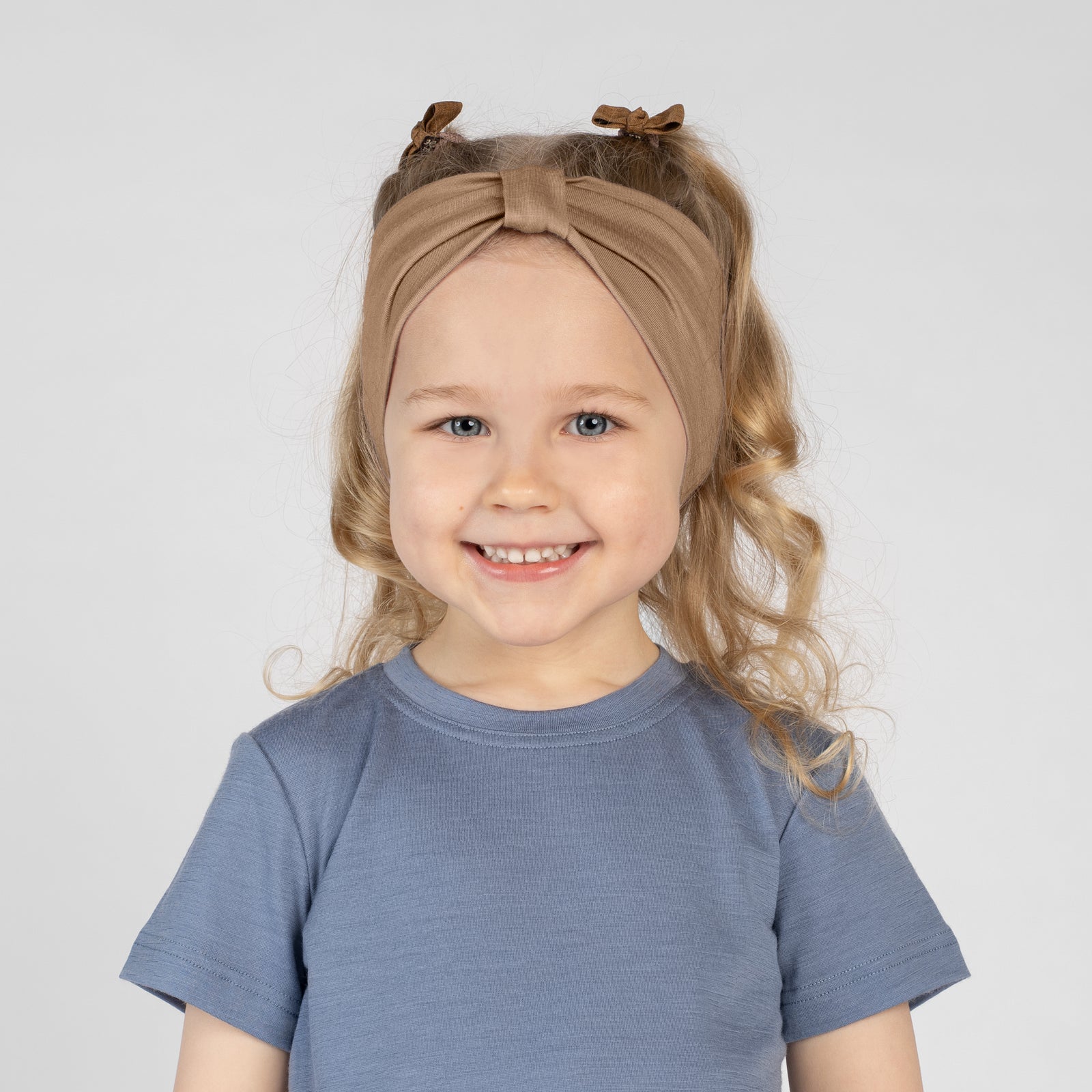 A close-up studio portrait of a young girl smiling directly at the camera against a plain light grey background. She is wearing a black short-sleeved t-shirt and a beige ribbon headband.