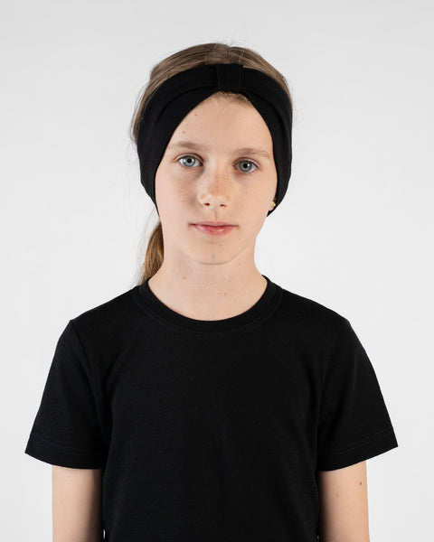A close-up studio portrait of a young girl looking directly at the camera with a neutral expression against a white background. She is wearing a black short-sleeved t-shirt and a black ribbon headband with a knot detail on top.