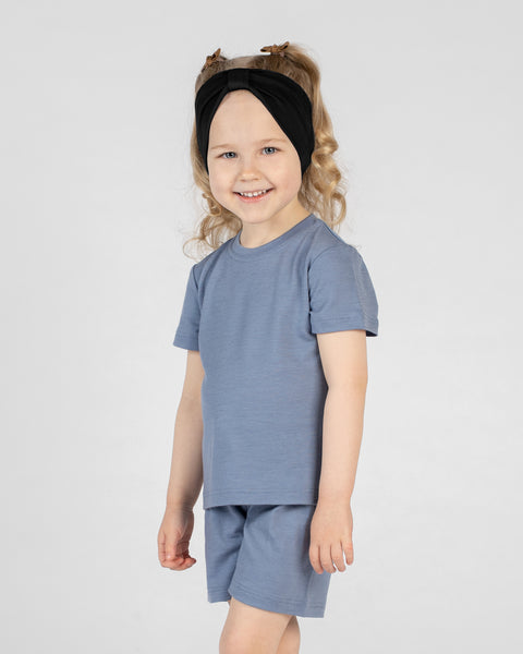 A studio portrait of a young, blonde-haired girl smiling brightly at the camera against a white background. She is wearing a dusty blue short-sleeved t-shirt and matching shorts, along with a black ribbon headband.