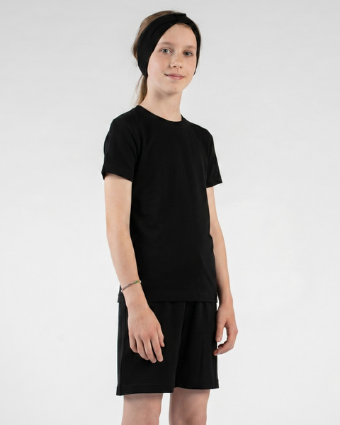 An older child stands against a light gray background wearing a black short-sleeve t-shirt, matching black shorts, and a knotted black headband. They are looking directly at the camera with a gentle smile, with their arms resting comfortably at their sides.