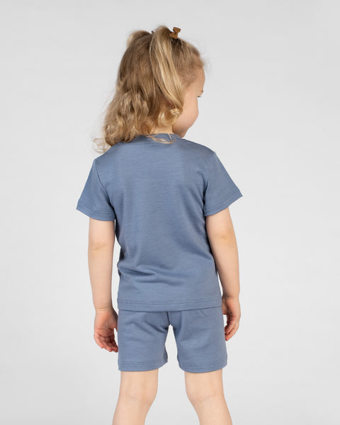 A child with blonde hair styled in a half-up ponytail stands facing away from the camera, wearing a dusty blue short-sleeve t-shirt and matching shorts. They are positioned against a plain light gray background.