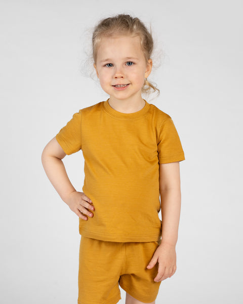 A child poses against a light gray background in a bright yellow short-sleeve t-shirt and matching shorts. They are smiling comfortably at the camera with one hand positioned on their hip.