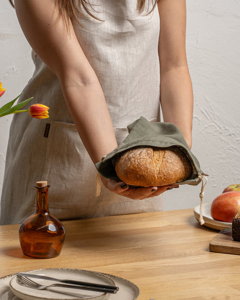 A person in a linen apron holds a loaf of bread on a wooden table with other food items.