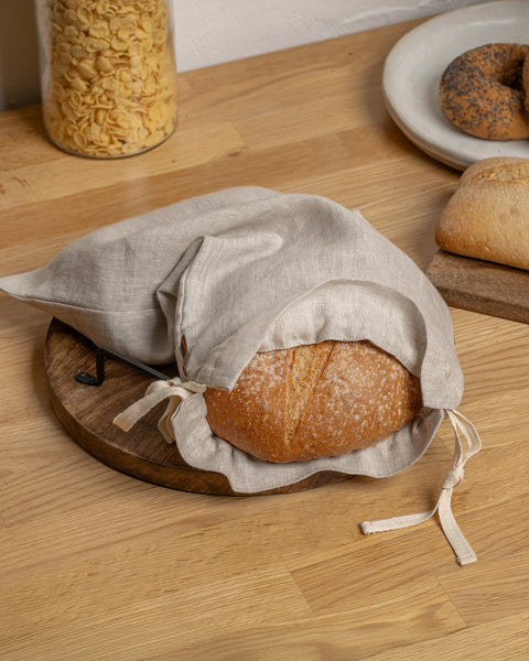 Loaf of bread in a cloth bag on a wooden surface with a plate of bagels in the background.