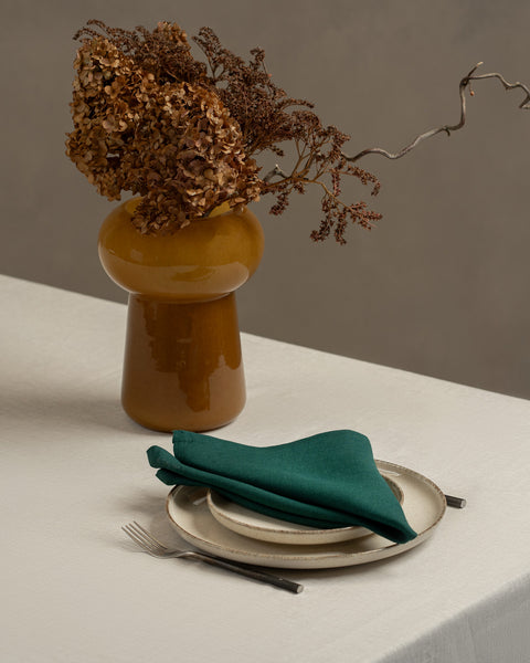 A tabletop covered with a linen tablecloth, with a vase and a single plate set on top of it. By the sides of the plates there are cutlery items, and there is a linen napkin in dark green color on top.