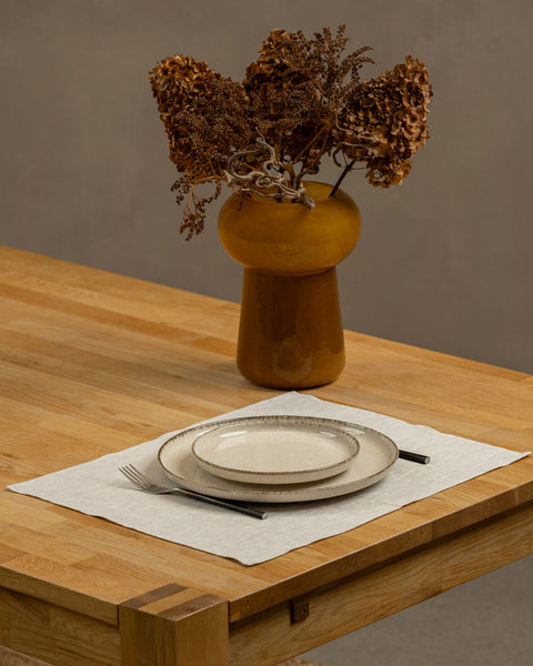 The menique linen placemat in natural color on a wooden table top. There is a vase on top of the table, as well as some cutlery by the sides of the plate, that is on top of the placemat.