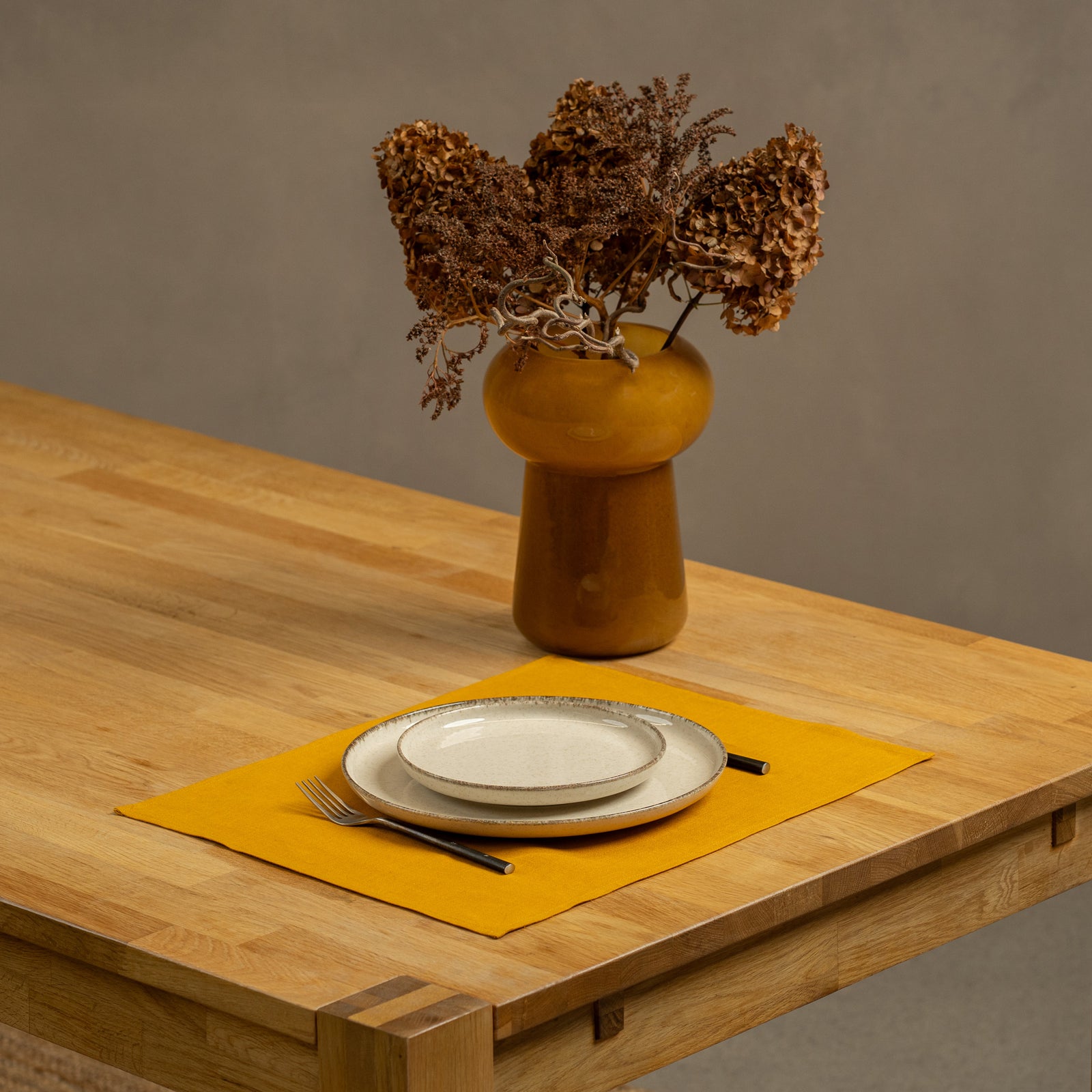 The menique linen placemat in dark green color on a wooden table top. There is a vase on top of the table, as well as some cutlery by the sides of the plate, that is on top of the placemat.