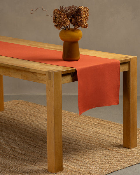 A wooden table positioned on top of a carpet. There is a cinnamon red linen table runner and a vase on top.