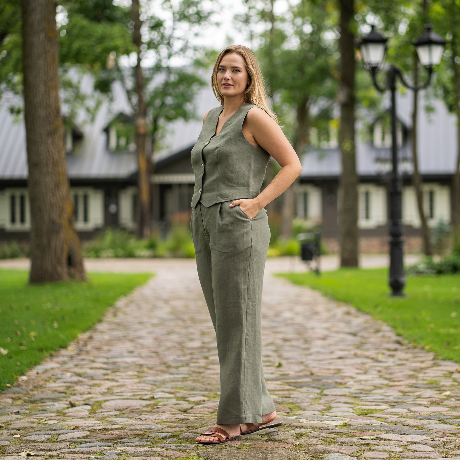 A person is shown from the waist down, modeling stone green, high-waisted, linen wide-leg trousers. They are wearing black open-toe sandals with thin straps and have their hands on their hips. The setting appears to be outdoors on an asphalt path, with a black metal fence and greenery visible in the background.