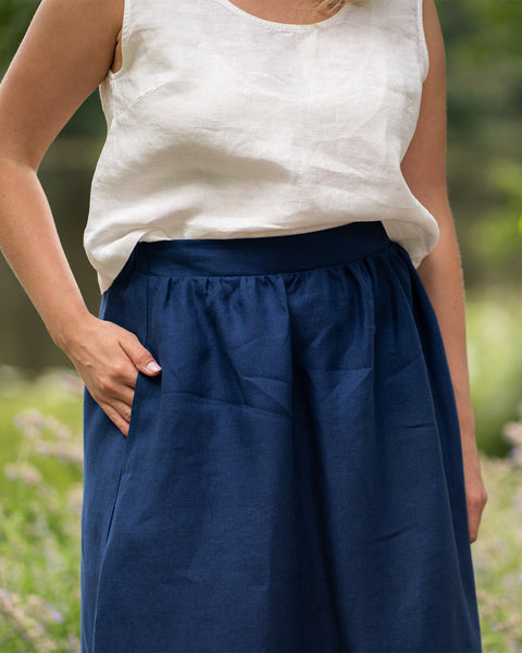 A close-up shot focuses on a woman's midsection as she stands outdoors, resting one hand comfortably inside the pocket of her skirt. The outfit features a relaxed-fit, pure white sleeveless linen top paired with a gathered storm blue midi skirt.