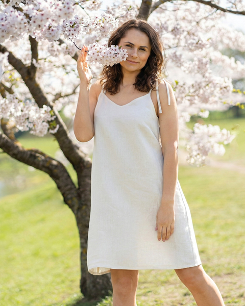Woman wearing pure white linen summer dress with adjustable tie straps, breathable relaxed-fit dress styled outdoors under cherry blossom trees