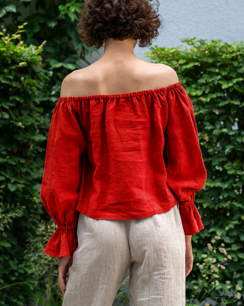 A woman with short, curly brown hair  standing in front of a green ivy-covered wall. She is wearing a pure red off-the-shoulder linen top with voluminous sleeves that gather at the wrists, and light-colored pants.