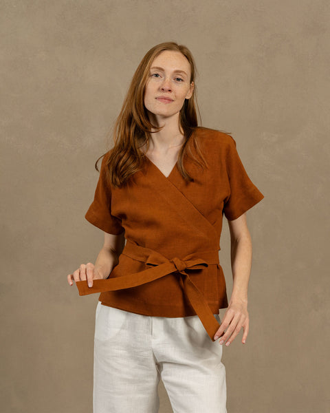 A fair-skinned person with long, auburn hair, wearing V-neck linen wrap top with short, wide sleeves and light-colored pants. They are holding the ends of the top's tie in front of them, forming a bow.