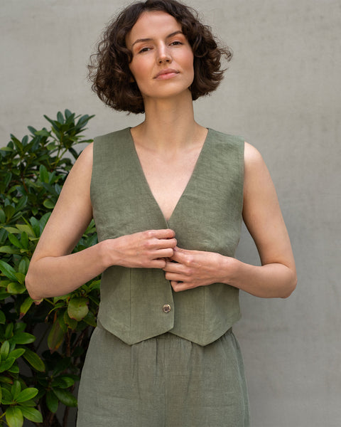 A woman with short, dark, curly hair stands facing forward, looking slightly upwards. She is wearing a sleeveless, stone green linen vest. Her hands are near the button, appearing to adjust or fasten it. She is also wearing matching stone green linen pants.