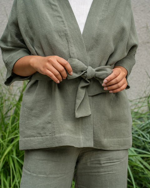 A person with light skin and dark hair stands outdoors, wearing a stone green linen kimono-style top and matching wide-leg pants. Their right hand is in the pocket of their pants. Tall green grasses and other plants are visible to their left.