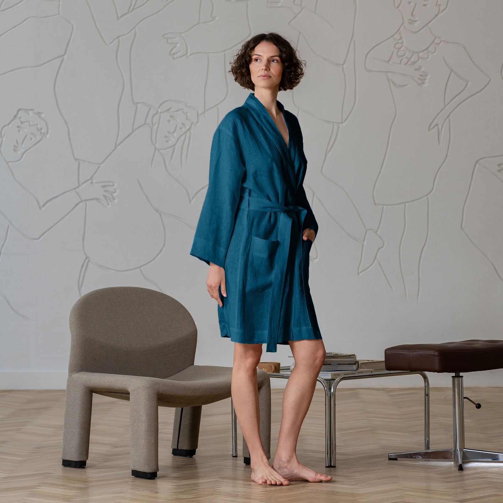 The menique women's linen bathrobe in cobalt blue color. The woman is standing in front of a gray wall with a wooden hanger on it. She has one her arms crossed across her chest.