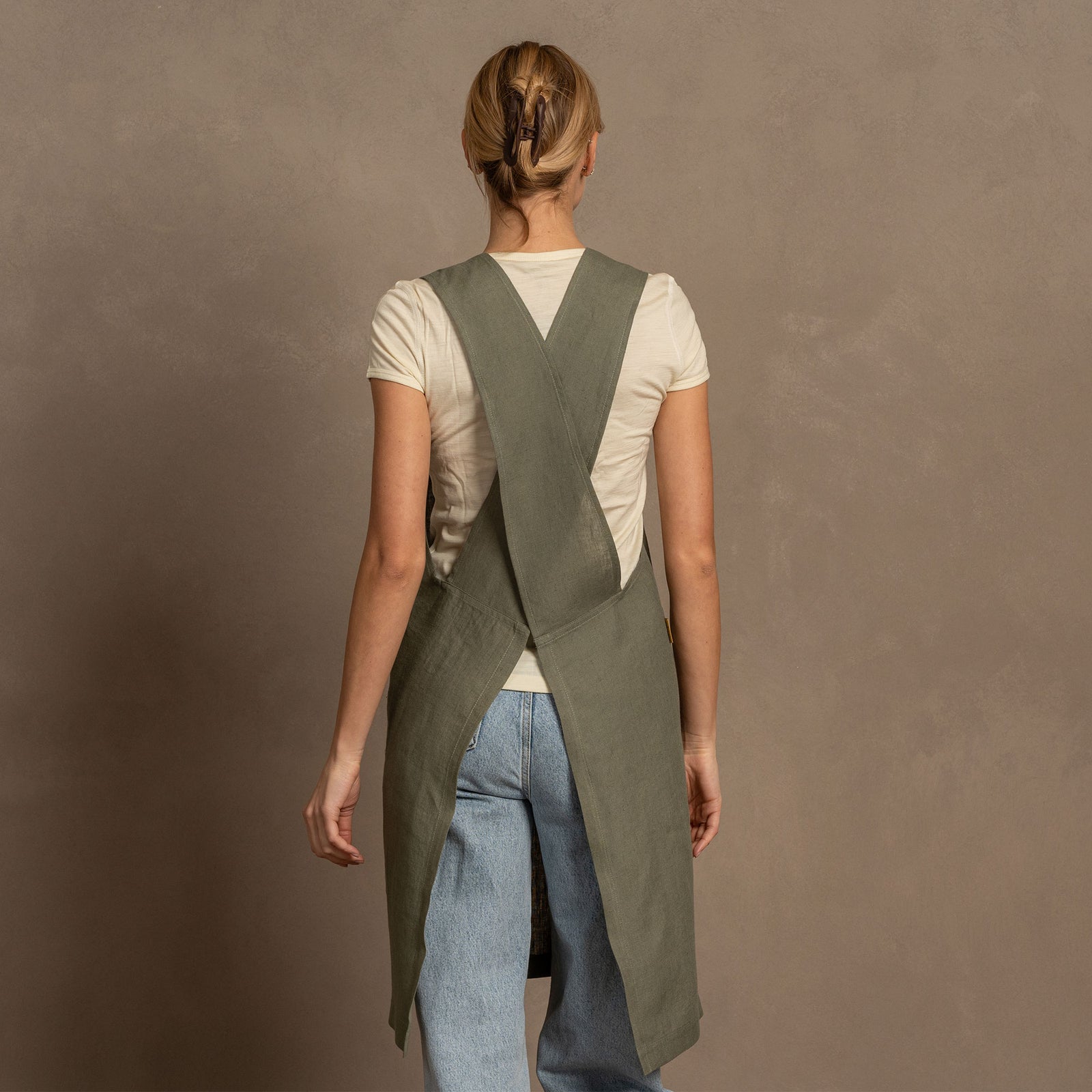 A woman with her hands in the pockets of a menique stone green linen cross-back apron, wearing a light blue jeans, stands smiling against a brown textured background.