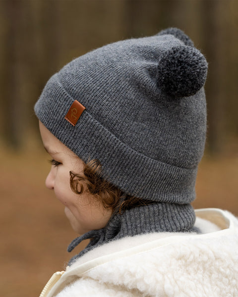 A child with light brown curly hair, is shown in profile, looking to the left. They are wearing a dark grey knit beanie with two dark grey pom-poms on top and a small brown label on the side, a grey turtleneck, and a cream-colored sherpa jacket. The background is a blurred outdoor setting with trees.