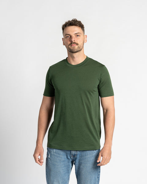 A man models a dark green crew neck merino wool t-shirt on a white background. The t-shirt is the focus of the image, showcasing its color and fit. He is also wearing light wash blue jeans.