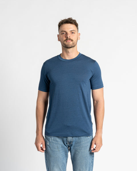 A man wearing a denim color 100% merino wool t-shirt and light blue jeans stands centered against a white background. The menique t-shirt is the primary focus, showcasing its regular fit and appearance. The man has short brown hair and a light beard, and is looking directly at the viewer.