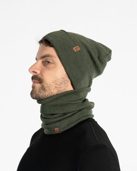 A man with a light brown beard is shown from the chest up, looking to the left of the frame. He is wearing a dark green knitted beanie and a matching neck gaiter. Both items of clothing have a small brown leather patch with a menique logo on them. The background is a plain white.