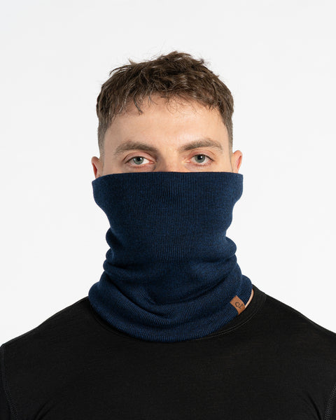 A person with short brown hair and light eyes is shown in a close-up studio shot against a white background. They are wearing a blue knit neck gaiter that covers their mouth and nose, and a black long-sleeved shirt. A small brown tag is visible on the neck gaiter. The person is looking directly at the camera.