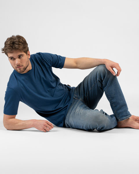 A man sitting on the floor, leaning back on one arm, wearing a blue short-sleeve t-shirt and blue jeans.