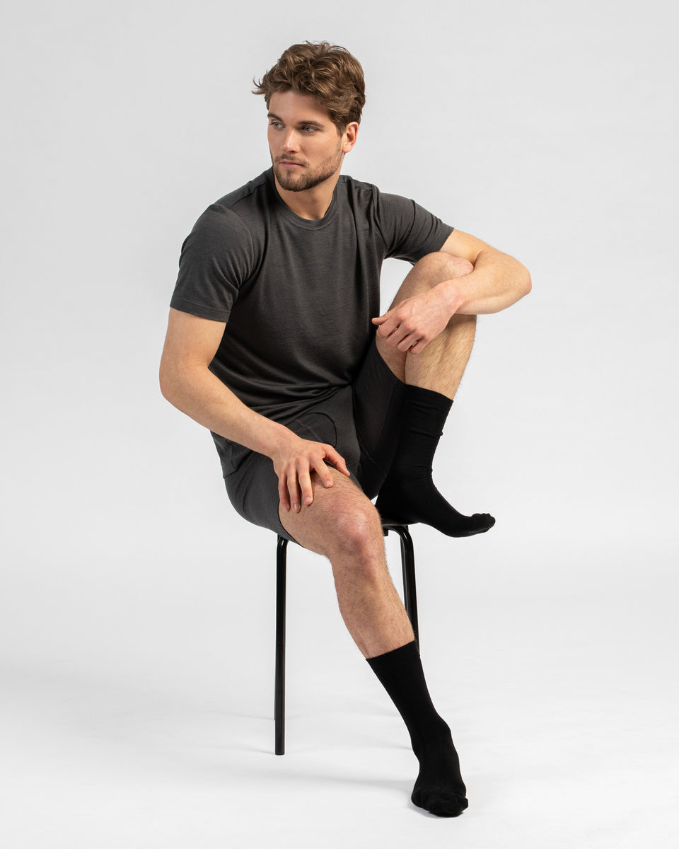 A studio portrait of a man sitting on a black stool, looking off to his right against a plain light grey background. He is wearing a matching set of charcoal grey short-sleeved t-shirt and long boxer briefs, paired with black crew socks.