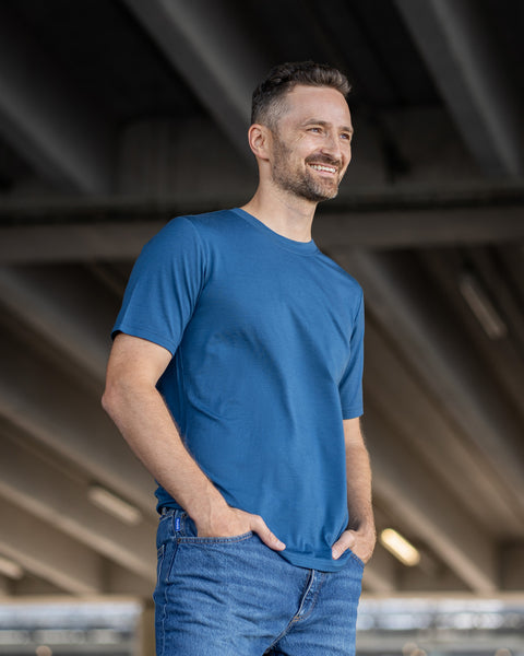 A smiling man with short dark hair and a beard stands with his hands in the front pockets of his blue jeans, looking off to the right. He is wearing a denim-blue short-sleeve crew neck t-shirt. The background is a shaded outdoor area under a concrete structure.