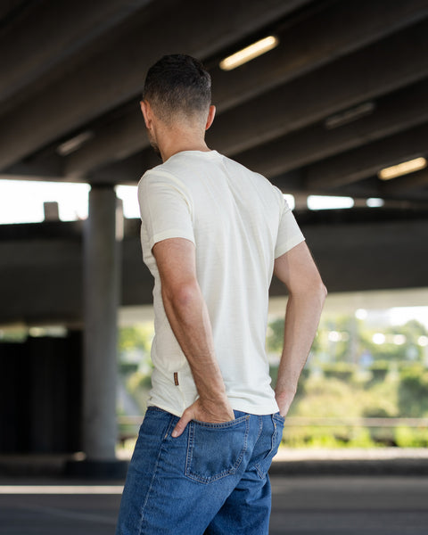 A man is photographed from behind, standing with both hands tucked into the back pockets of his blue jeans. He is wearing an off-white, short-sleeve t-shirt that features a small brown logo tag near the bottom left hem. The setting is under a concrete bridge or overpass.