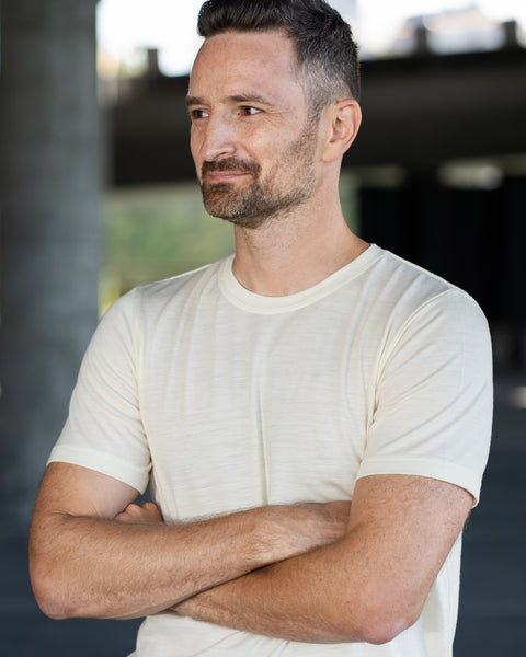 A man with short dark hair and a beard stands outdoors with his arms crossed, looking thoughtfully to his right with a slight smile. He is wearing an off-white, natural-colored short-sleeve crew neck t-shirt. The background consists of blurred concrete pillars under an overpass.