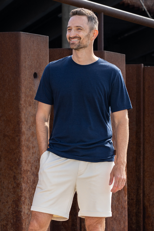 A smiling man standing outdoors in front of rusty metal pillars. He is wearing a navy blue short-sleeve crew neck t-shirt and beige shorts, with his right hand casually resting in his pocket as he looks off into the distance.