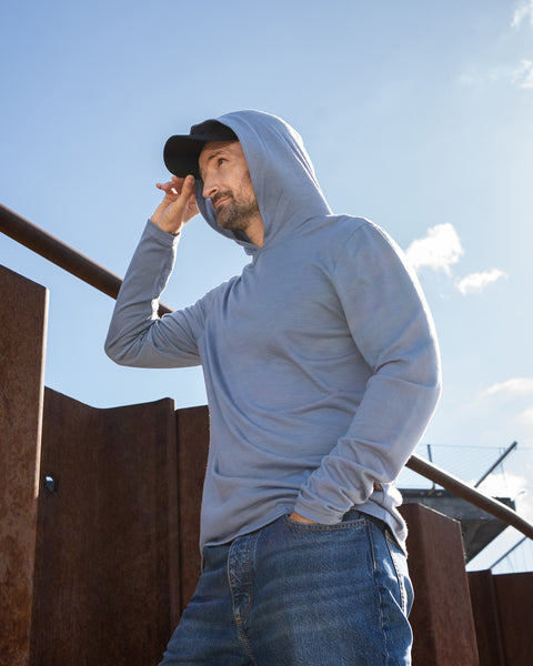 A man stands outdoors on a sunny day wearing a dusty blue hoodie with the hood pulled up over a black baseball cap. He has one hand in his jeans pocket and the other lightly touching the brim of his cap. A rusty corrugated metal barrier and a concrete bridge are visible in the background.